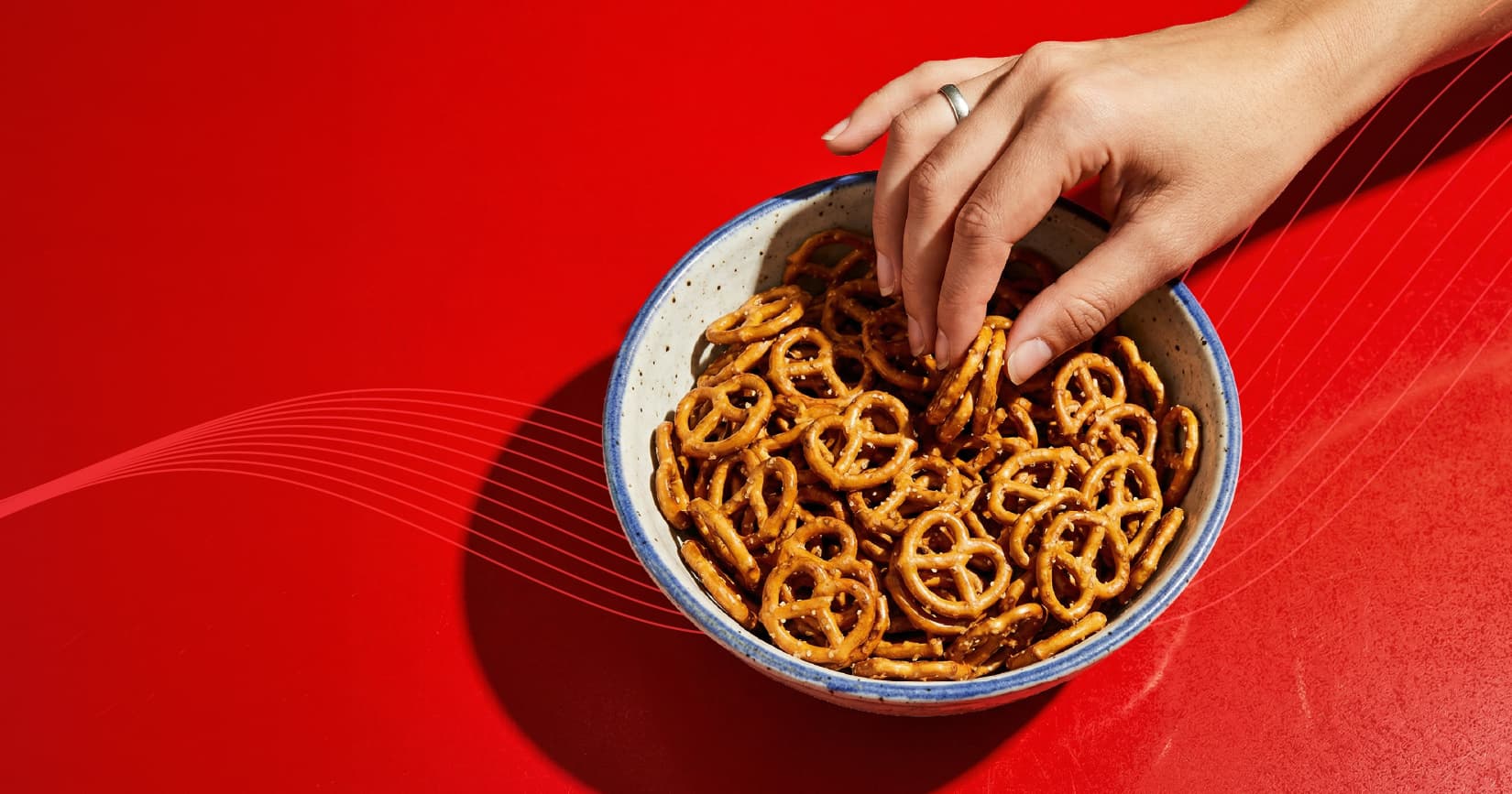 A woman's hand reaching towards a bowl of pretzels set against a red background.