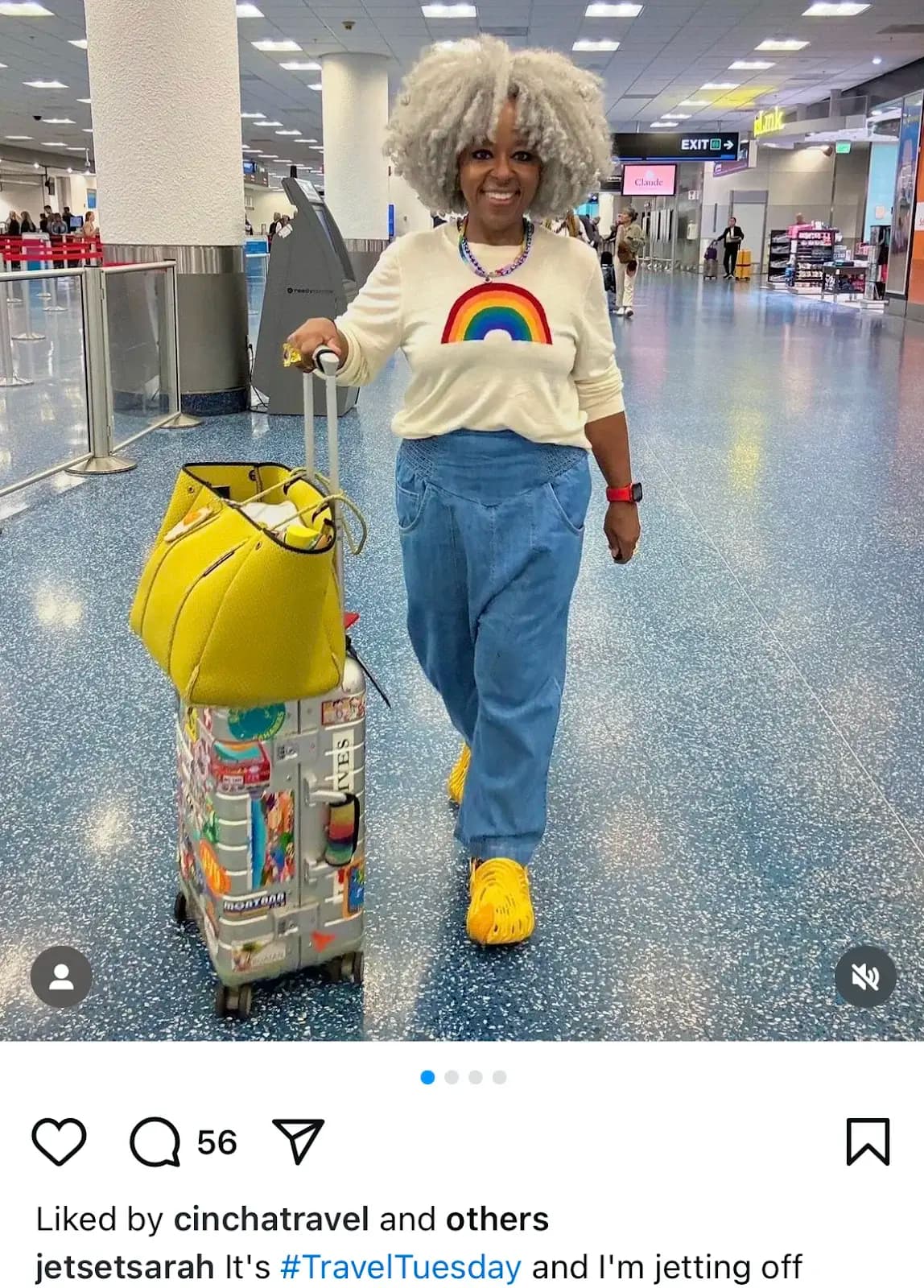 A woman with gray curly hair, wearing a rainbow sweater and blue jeans, walks through an airport with luggage and a yellow bag.
